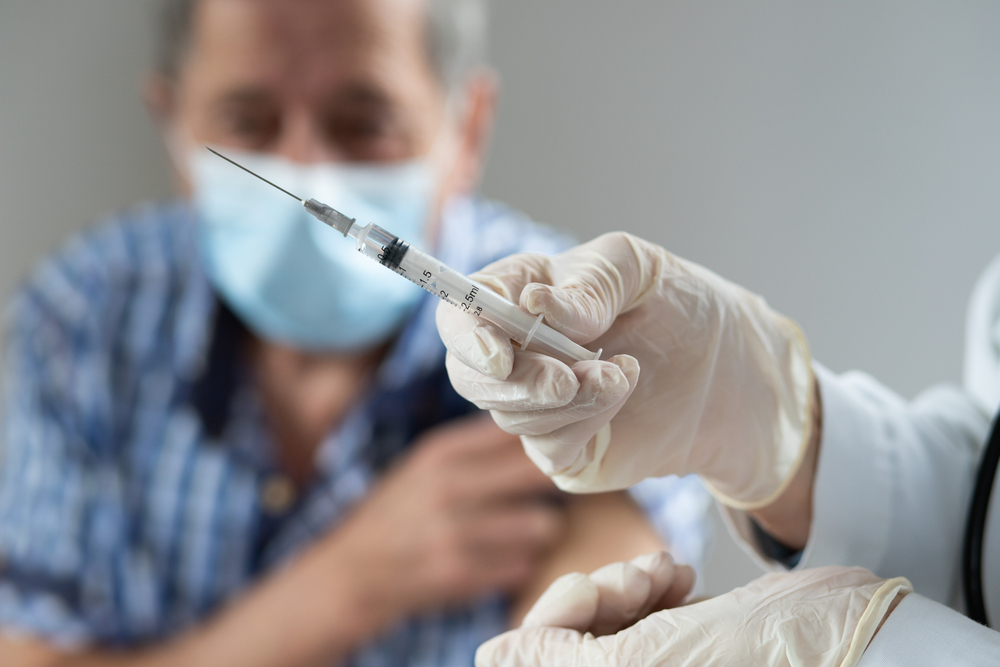 a needle with the COVID-19 vaccine in it and a senior man in the background waiting to receive his dose