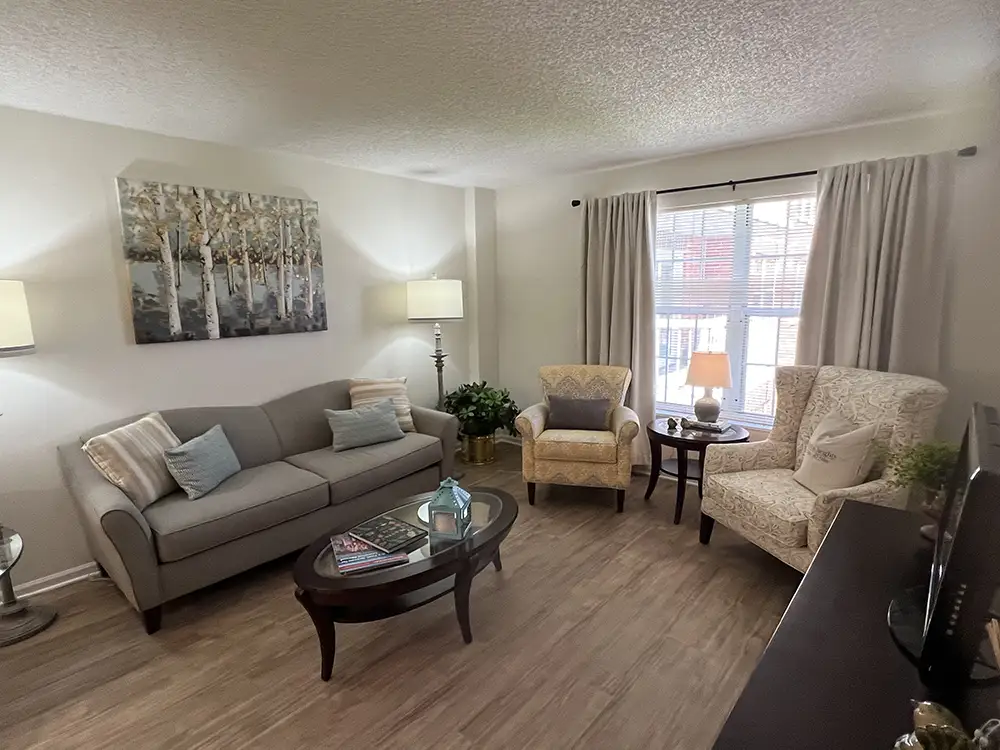 A brightly lit living room in a senior apartment at The Moorings of Arlington Heights, furnished with a gray sofa, two accent armchairs, an oval coffee table, and large window curtains over hardwood-style flooring.