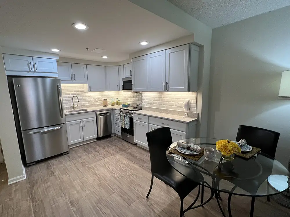 A modern senior living apartment kitchen and dining area at The Moorings of Arlington Heights, featuring white cabinetry, subway tile backsplash, stainless steel appliances, and a glass-top dining table set for two.