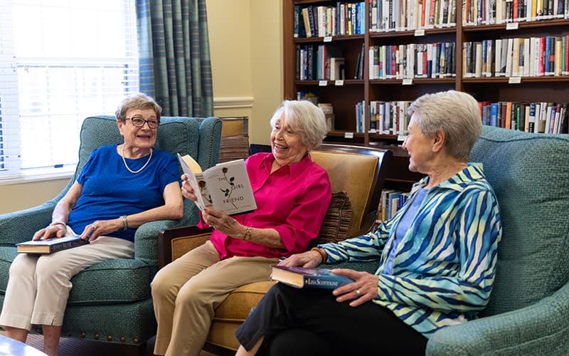 Three senior women reading books in the library at The Moorings of Arlington Heights