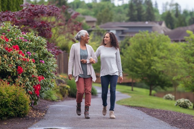 a senior woman and her adult granddaughter strolling through a garden with their arms linked