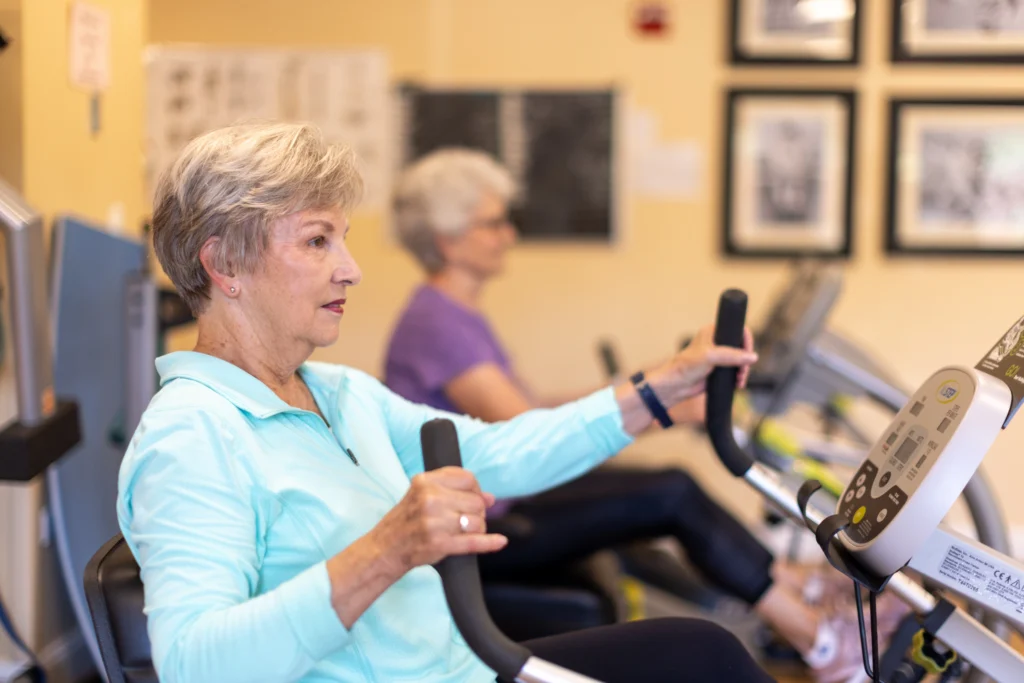 senior woman in a bright light blue quarter zip sweatshirt works out her arms on a piece of equipment in the fitness center of an independent living community for seniors