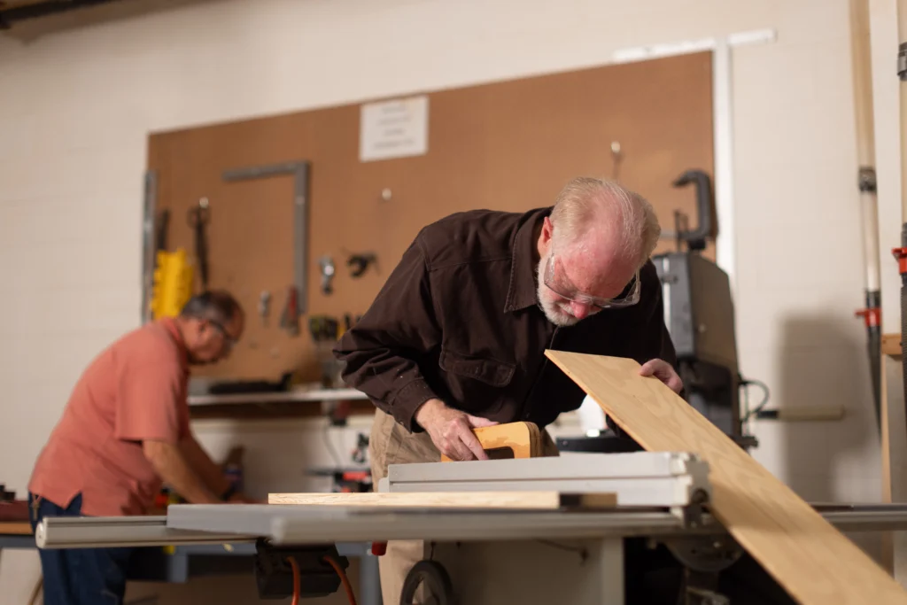 senior man leans over a workbench in a woodworking shop in an independent living community, working on a wood project