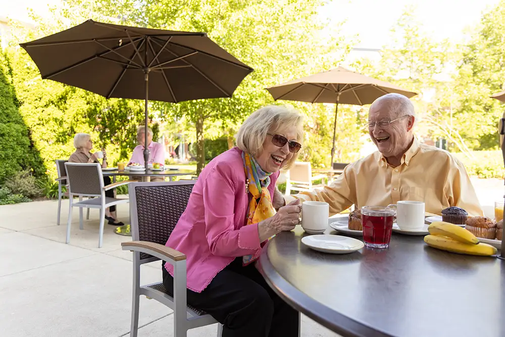 Lake Forest Place residents laugh together over coffee and snacks at an outdoor patio table shaded by an umbrella, embracing relaxed cottage living.