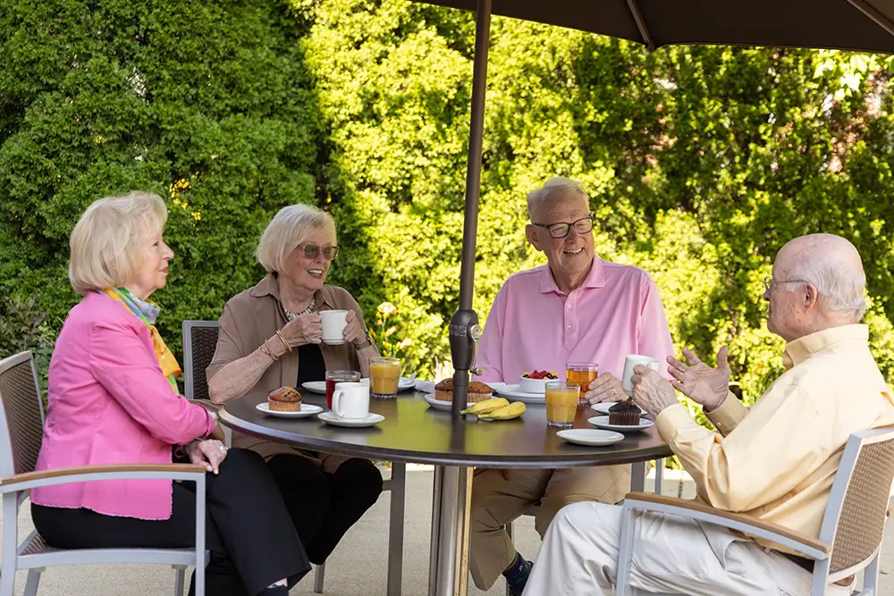 Four seniors at Lake Forest Place sit around an outdoor patio table under an umbrella, smiling and chatting over coffee, juices, and pastries on a sunny day