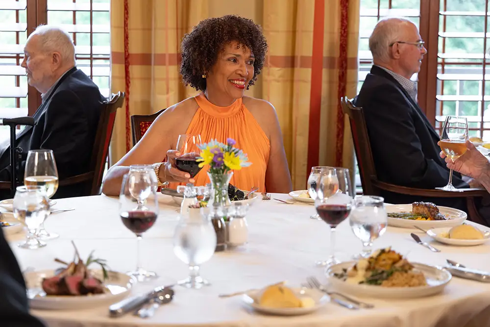 A Lake Forest Place resident in an orange dress smiles warmly at a set dining table, holding a glass of red wine during a group dinner with other residents.