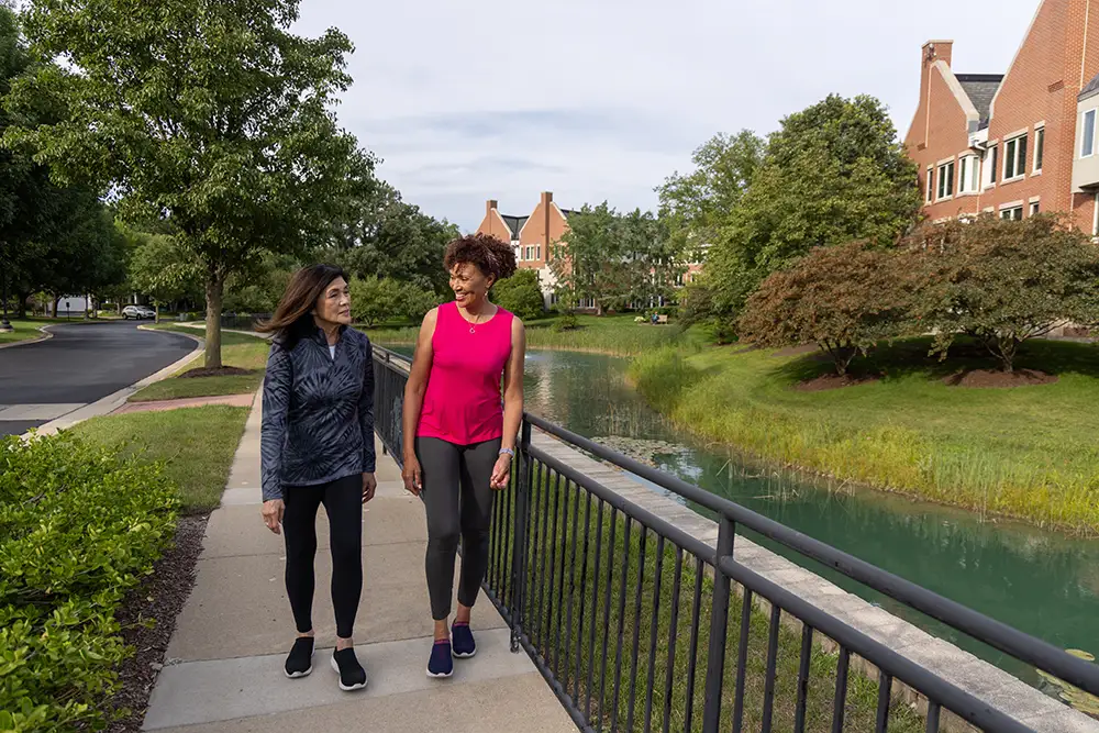 Two seniors at Lake Forest Place take a refreshing walk along a paved sidewalk next to a scenic pond and surrounding brick homes, enjoying active cottage living.