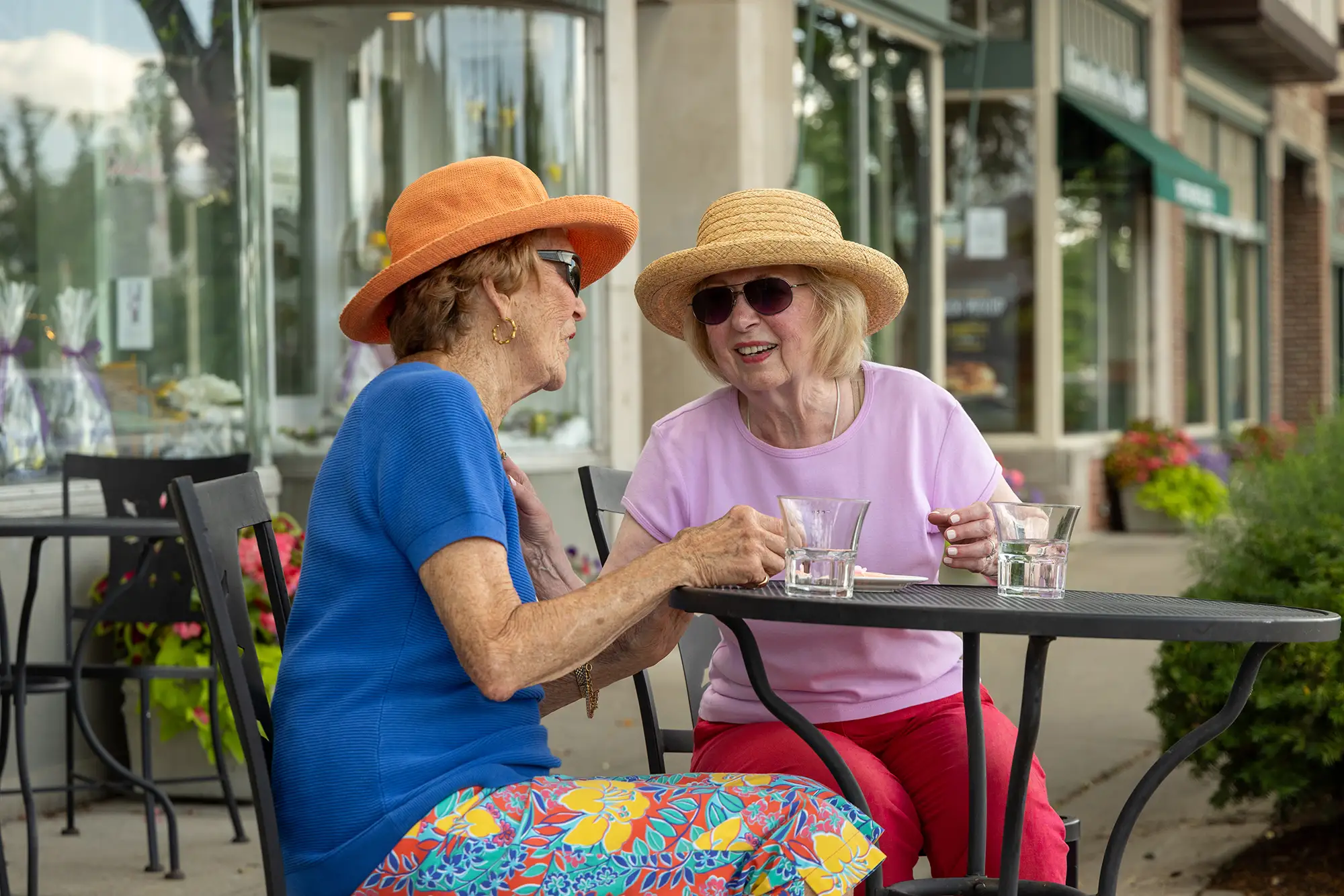 Two seniors at Lake Forest Place wearing sun hats sit at an outdoor cafe table along a storefront sidewalk, talking and laughing together over glasses of water.