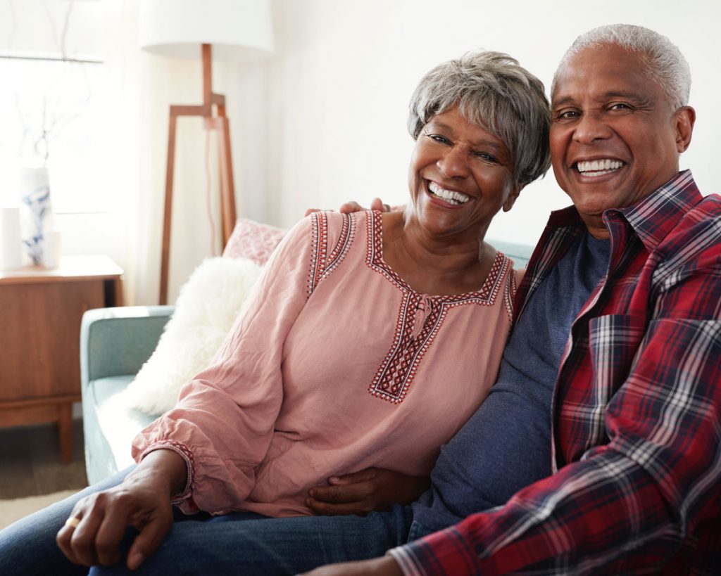 an elderly couple smiling and sitting on a couch inside of their senior apartment near chicago