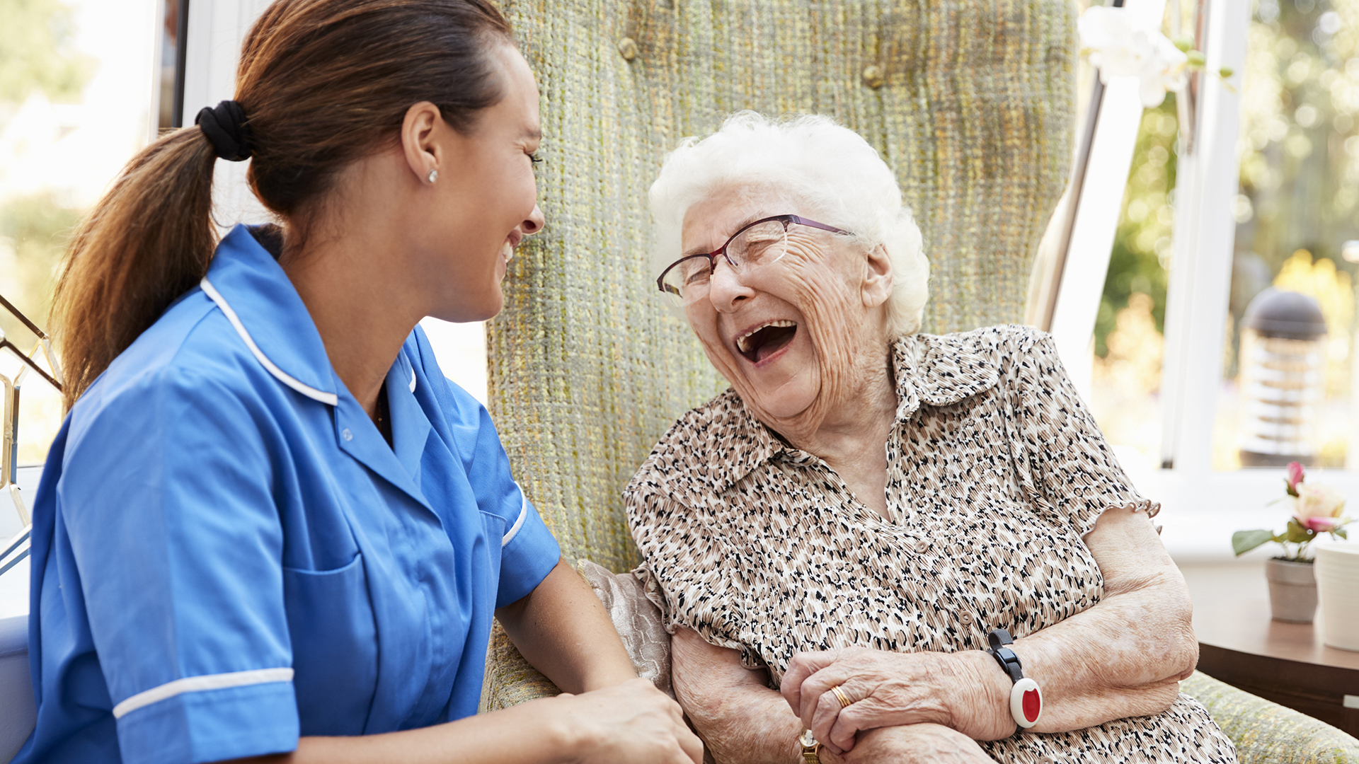 Senior Woman Sitting In Chair And Laughing With Nurse In Retirement Home Senior Woman Sitting In Chair And Laughing With Nurse In Retirement Home