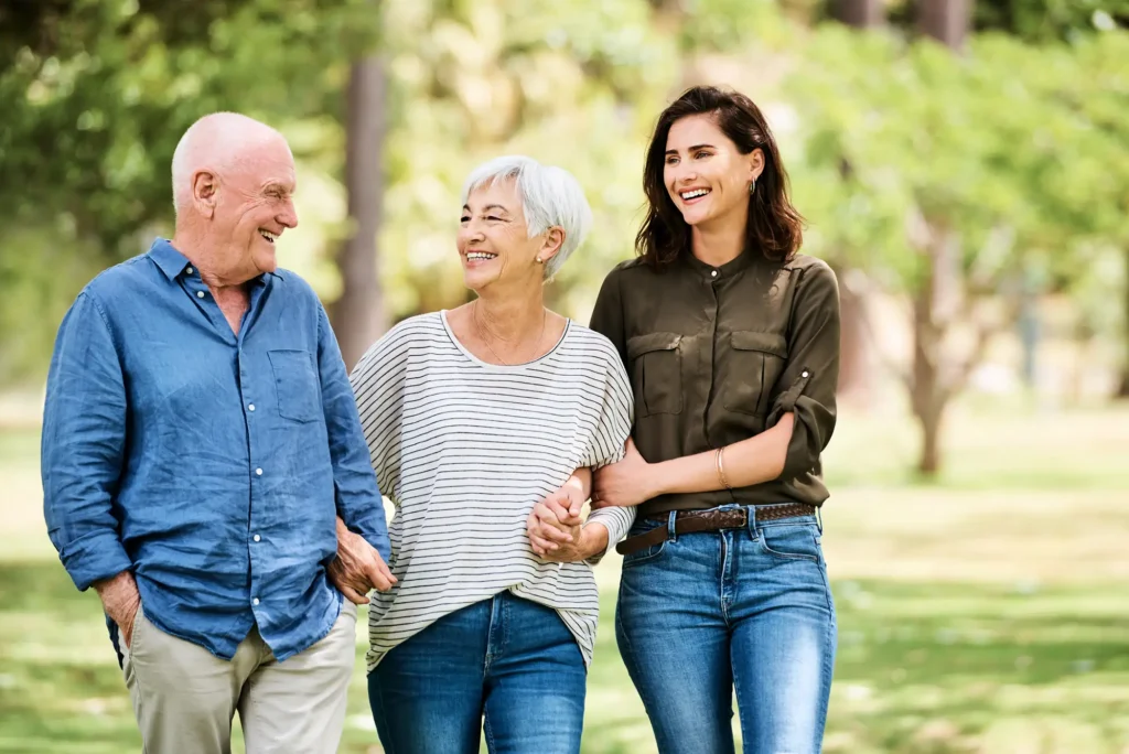 An adult daughter walks arm-in-arm through a park setting while visiting her parents at Westminster Place, smiling and sharing a happy moment outdoors.