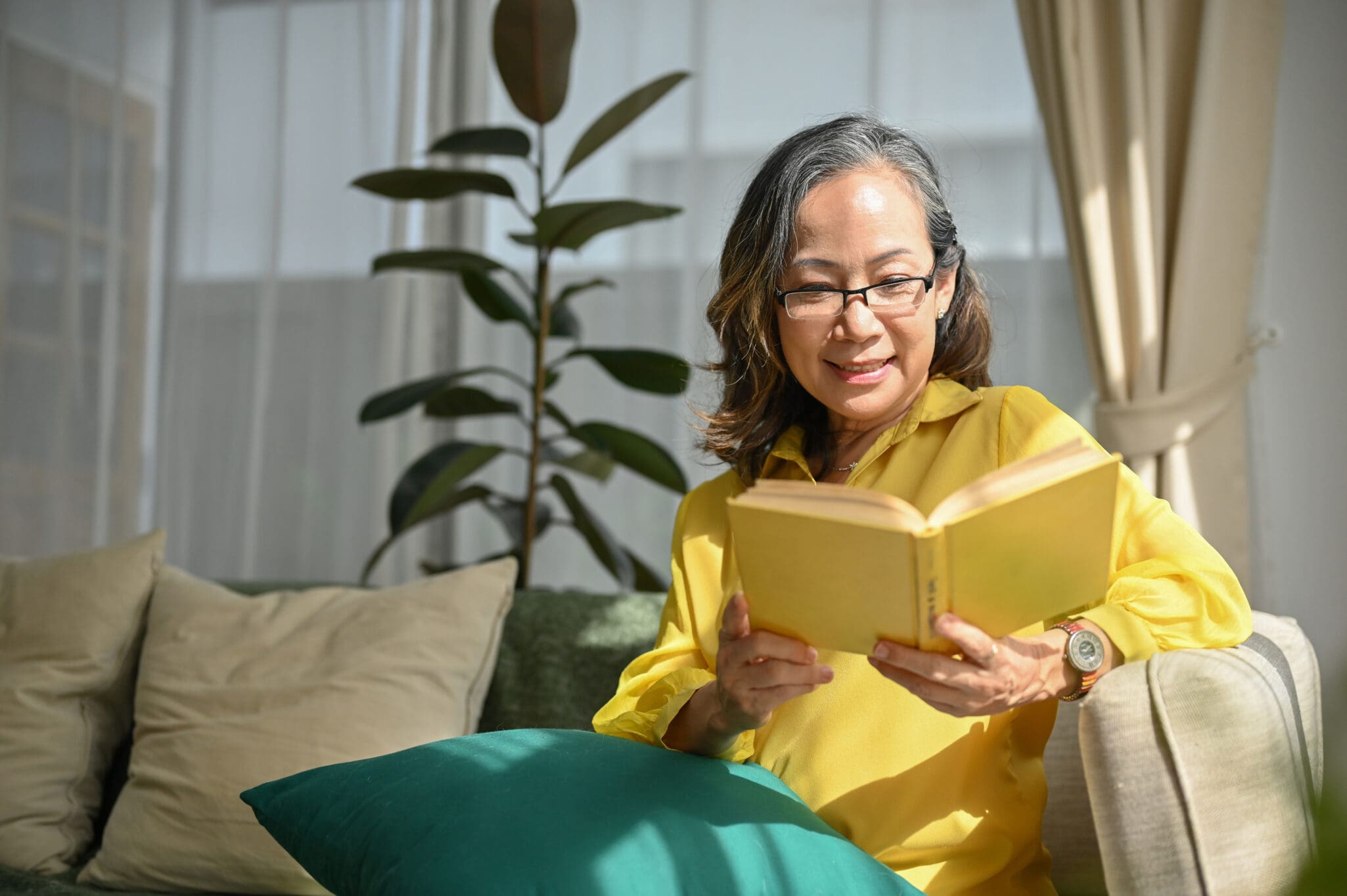 Relaxed asian aged-woman in glasses sitting and reading a book in comfortable living room. woman reading a book