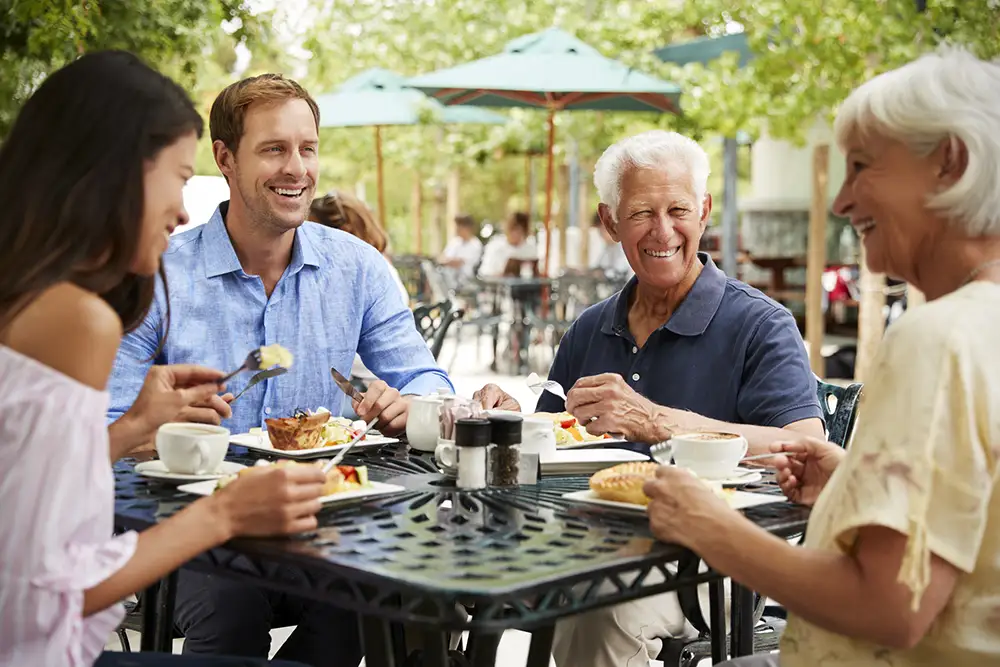 An adult son and daughter enjoy an outdoor patio lunch while visiting their aging parents at Westminster Place, all laughing and eating together around a black wrought-iron table.
