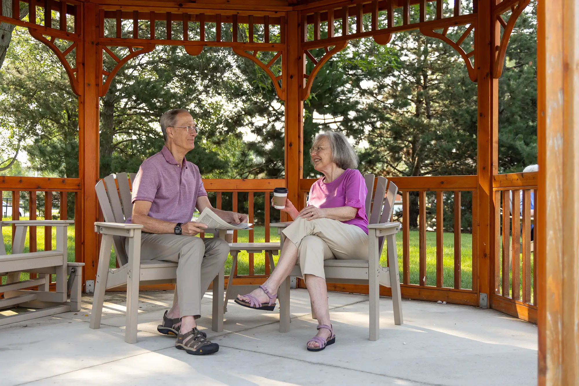 Two seniors at The Moorings of Arlington Heights are sitting and chatting in a wooden gazebo, looking relaxed in comfortable chairs.