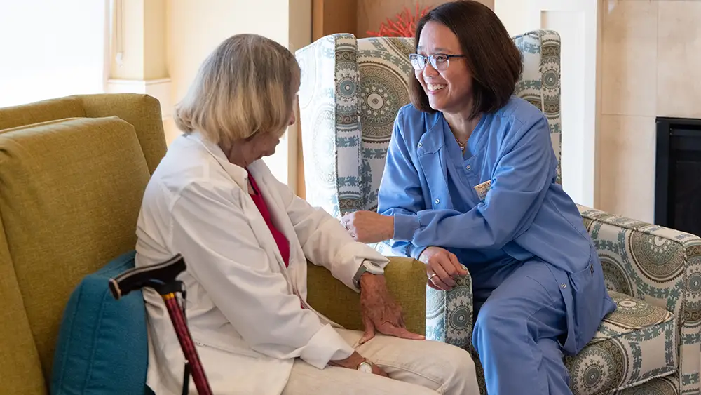 Skilled Nursing patient sitting on a sofa and speaking with her caregiver at The Moorings of Arlington Heights