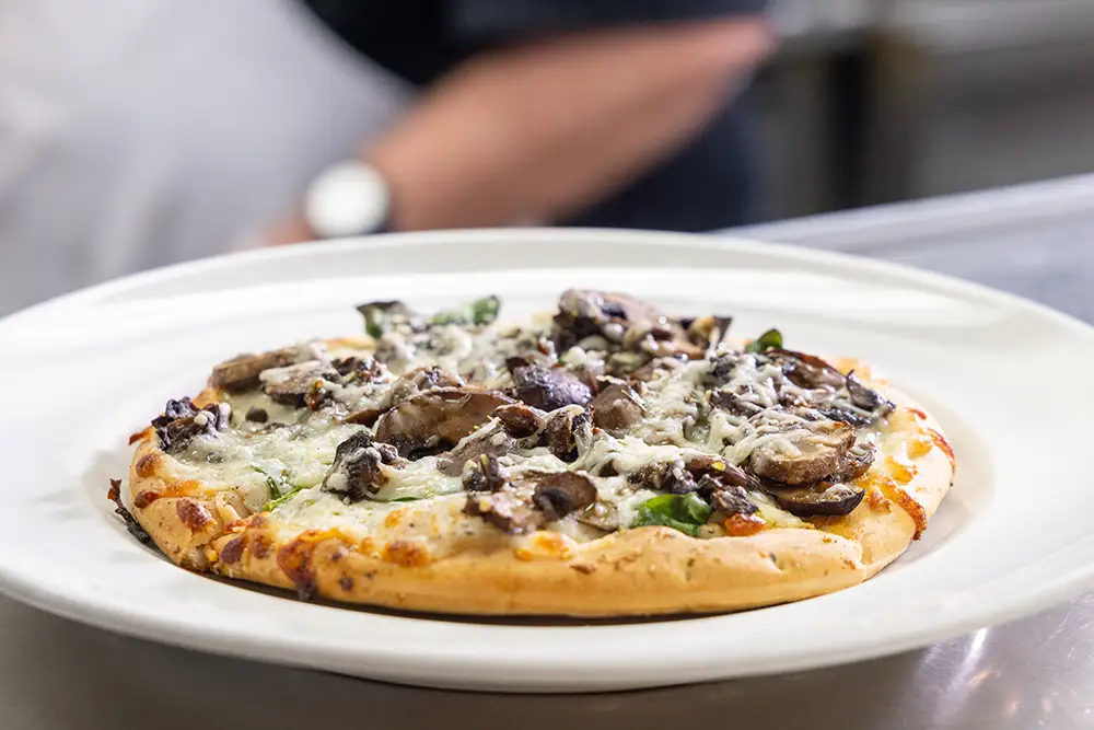 A freshly baked pizza topped with melted cheese, spinach, and sautéed mushrooms, served on a white plate for The Moorings of Arlington Heights residents to enjoy.