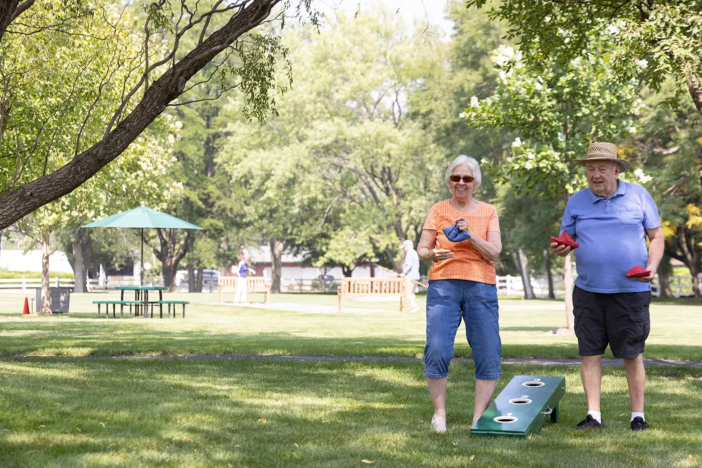 Two seniors at The Moorings of Arlington Heights play a game of cornhole outdoors on a grassy lawn surrounded by green trees during a bright autumn afternoon.
