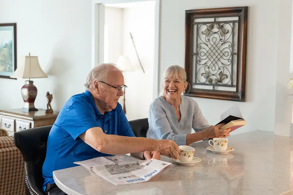 A senior resident at The Moorings of Arlington Heights reads a newspaper at his kitchen counter, holding a cup of coffee. Another senior resident is smiling, reading a red book next to him.